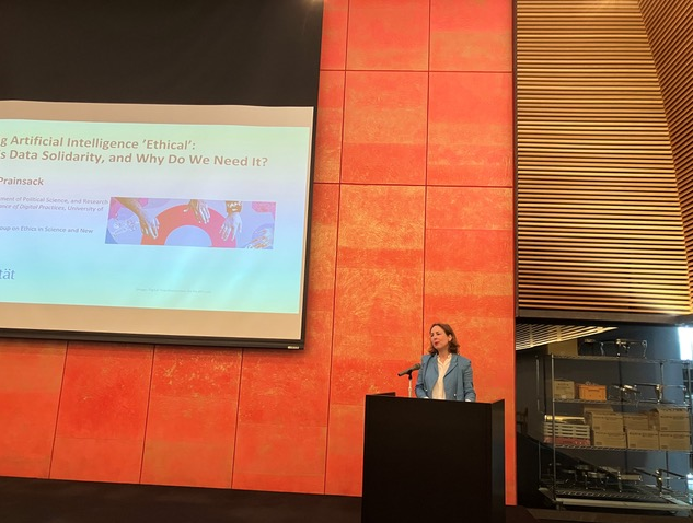 Prof Barbara Prainsack standing behind a lectern during her keynote with presentation slides shown on a screen on the left side behind her. 